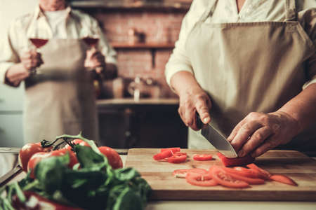Cropped image of beautiful senior couple in aprons drinking wine and chopping tomato while cooking together in kitchenの写真素材