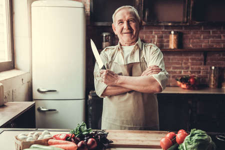 Handsome senior man in apron is holding a knife, looking at camera and smiling while cooking in kitchenの写真素材