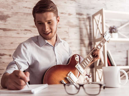 Handsome young businessman is playing guitar, making notes and smiling in his office during the breakの写真素材