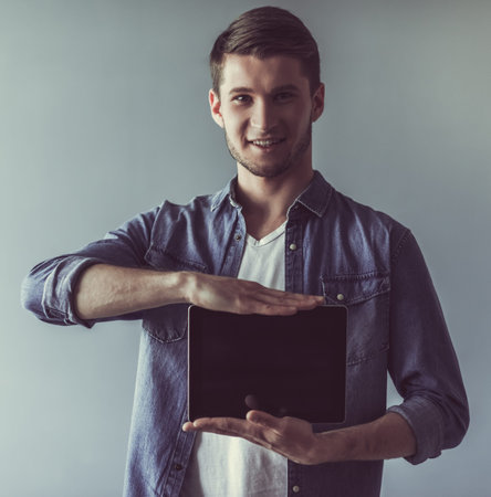 Handsome guy in casual clothes is showing a digital tablet, looking at camera and smiling, on gray backgroundの写真素材