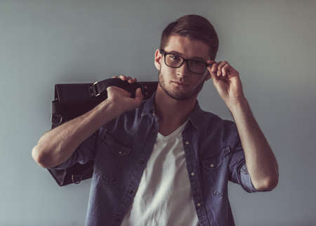 Handsome young man in casual clothes and glasses is holding a stylish bag, looking at camera and smiling, on gray backgroundの写真素材