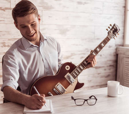 Handsome young businessman is playing guitar, making notes and smiling in his office during the breakの写真素材