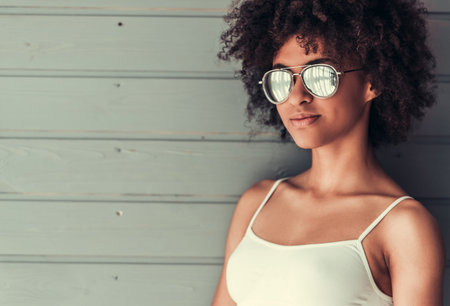 Beautiful Afro American girl in white top and sun glasses is looking at camera and smiling, on gray wall backgroundの写真素材