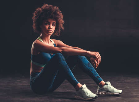 Attractive Afro American girl in sportswear is looking at camera while sitting on black backgroundの写真素材