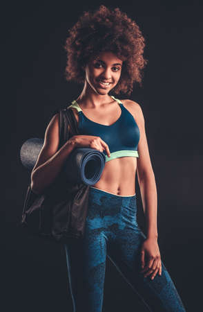 Attractive Afro American girl in sportswear is holding a yoga mat, looking at camera and smiling, on black backgroundの写真素材