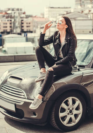 Beautiful girl in leather jacket and sun glasses is smoking while sitting on the hood of her carの写真素材