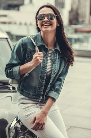 Beautiful girl in jean jacket and sun glasses is looking at camera and smiling while leaning on her carの写真素材