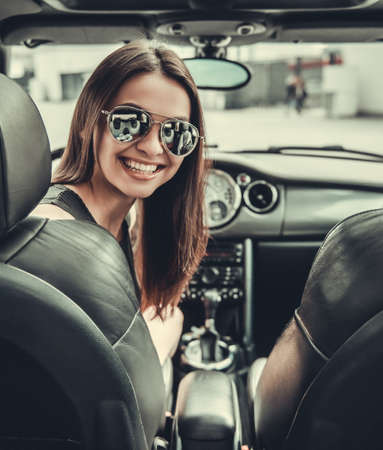 Beautiful girl in sun glasses is looking at camera and smiling while sitting in her carの写真素材
