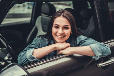 Beautiful girl in jean jacket is looking at camera and smiling while sitting in her carの写真素材
