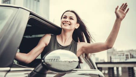 Beautiful girl is sticking out the window, waving and smiling while driving a carの写真素材