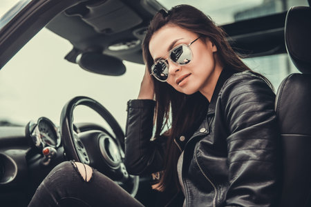 Beautiful girl in leather jacket and sun glasses is looking at camera while sitting in her car