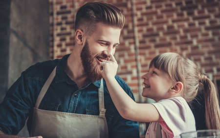 Cute little girl and her handsome bearded dad in aprons are having fun while cooking in kitchenの写真素材