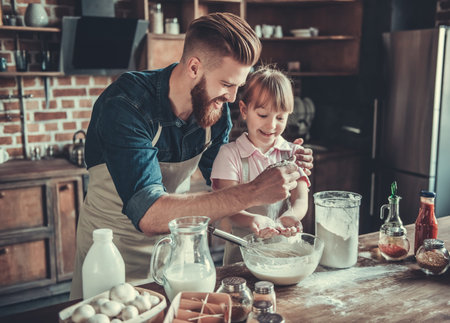 Cute little girl and her handsome bearded dad in aprons are preparing dough, adding flour and...
