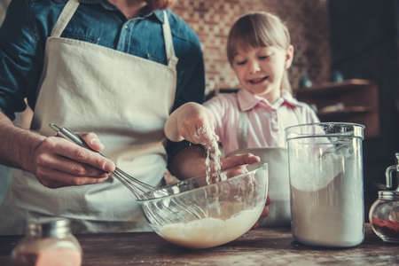 Cute little girl and her handsome bearded dad in aprons are whisking eggs, adding flour and smiling while cooking in kitchenの写真素材