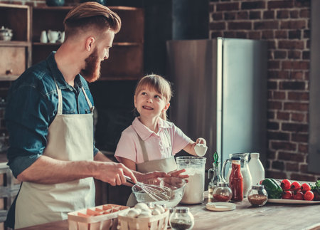 Cute little girl and her handsome bearded dad in aprons are whisking eggs and smiling while cooking in kitchenの写真素材
