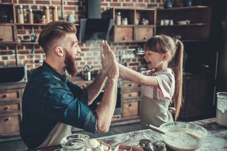 Cute little girl and her handsome bearded dad in aprons are having fun with flour, giving high five and smiling while cooking in kitchenの写真素材