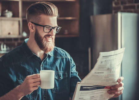 Handsome bearded young man is drinking coffee, reading a newspaper and smiling while resting in kitchenの写真素材