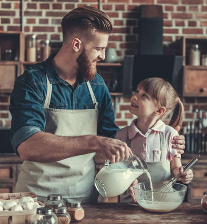 Cute little girl and her handsome bearded dad in aprons are preparing dough and smiling while cooking in kitchenの写真素材
