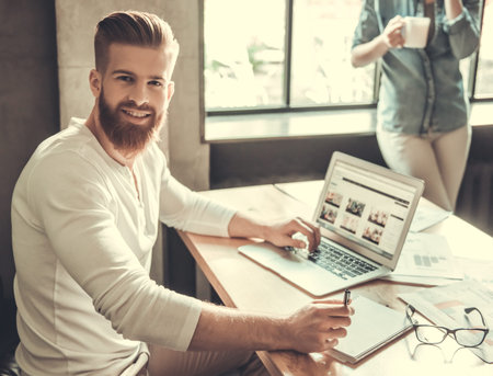 Young bearded man using a laptop while working in an office on the background of a colleague...