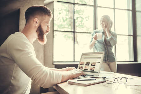 Young bearded man using a laptop while working in an office on the background of a colleague drinking tea near the window.の写真素材