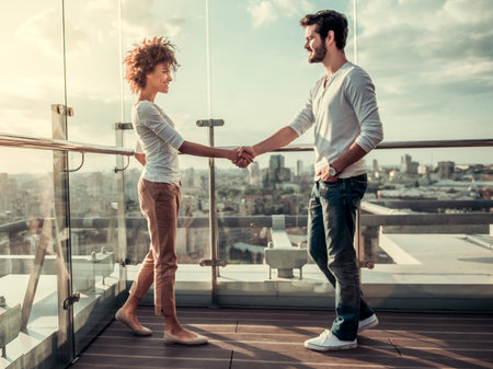 Beautiful young couple is shaking hands and smiling while standing on balcony of the city buildingの写真素材