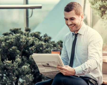 Handsome businessman is reading newspaper, drinking coffee and smiling while sitting outdoorsの写真素材