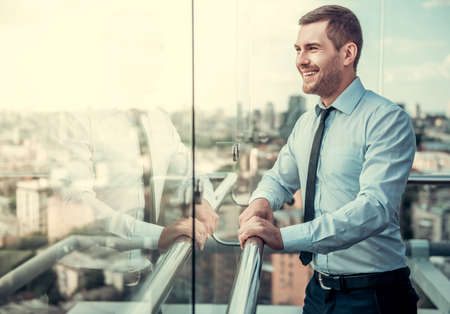Handsome businessman is looking out the window and smiling while resting on balcony of the office buildingの写真素材