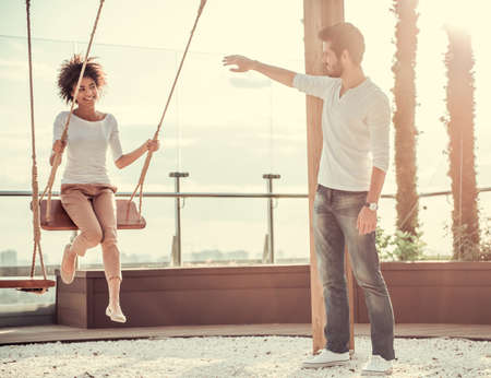 Beautiful young couple is talking and smiling while resting outdoors, girl is swingingの写真素材