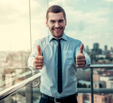 Handsome businessman is showing Ok sign, looking at camera and smiling while standing on balcony of the office buildingの写真素材