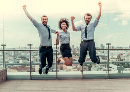 Beautiful business people are looking at camera and smiling while jumping on balcony of the office buildingの写真素材