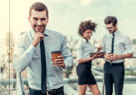 Handsome businessman with coffee on the foreground. Business people are communicating on balcony of the office building during coffee breakの写真素材