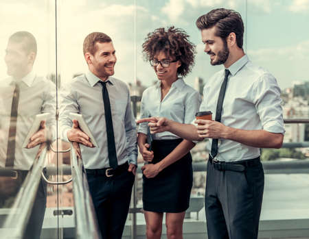 Beautiful business people are using a digital tablet, talking and smiling while standing on balcony of the office building during coffee breakの写真素材