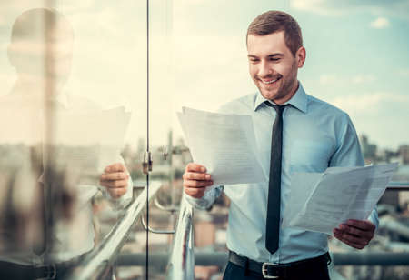 Handsome businessman is studying documents and smiling while standing on balcony of the office buildingの写真素材
