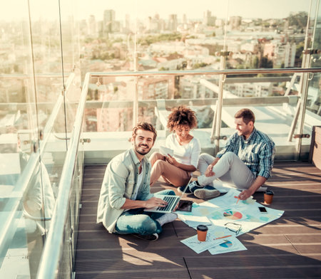 Beautiful young people are discussing projects, using gadgets and drinking coffee while working on balcony of the city buildingの写真素材
