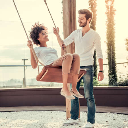 Beautiful young couple is talking and smiling while resting outdoors, girl is swingingの写真素材