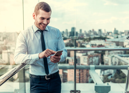 Handsome businessman is using a smart phone and smiling while resting on balcony of the office buildingの写真素材