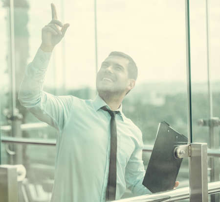Businessman is holding a folder, pointing upward and smiling while standing on balcony of the office building, view through the windowの写真素材