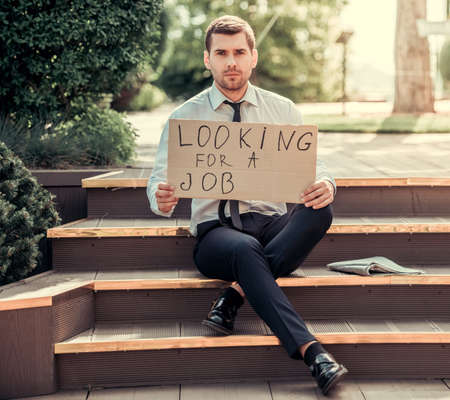 Handsome businessman is holding a cardboard sheet with phrase "looking for a job" and looking at camera while sitting on stairs outdoorsの写真素材