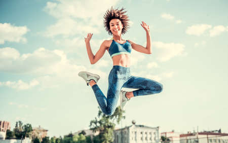 Beautiful Afro American girl in sportswear is looking at camera and smiling while jumping on city backgroundの写真素材