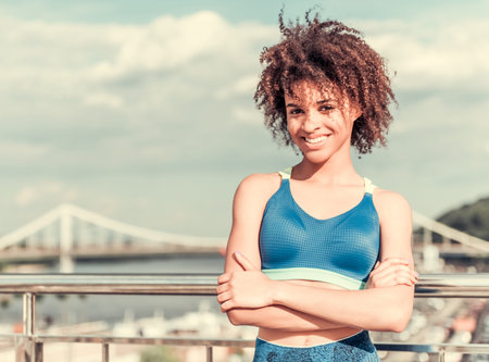 Beautiful Afro American girl in sportswear is looking at camera and smiling while resting on the city bridge during morning runの写真素材