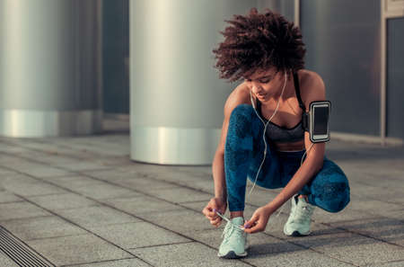 Beautiful Afro American girl in sportswear and earphones is lacing running shoes during morning runの写真素材