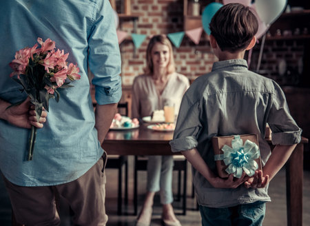 Family celebrating mother's birthday in decorated kitchen. Husband with flowers, son with a gift box, mother is smiling waiting for presentsの写真素材