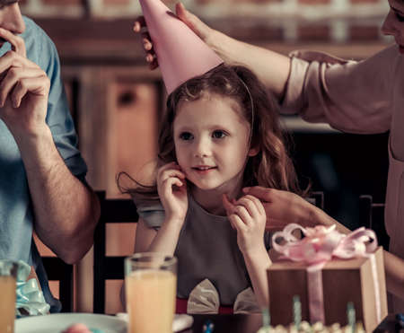 Happy family is sitting at the table in decorated kitchen during birthday celebration, mom is putting birthday hat on her cute daughterの写真素材