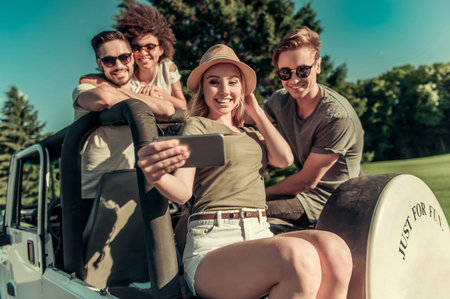 Happy young people are taking photo using a smart phone and smiling while travelling by car in sunny...