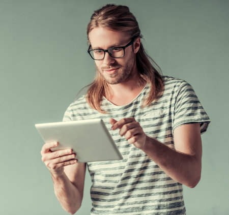 Stylish young man with shoulder-length blond hair and in eyglasses is using a digital tablet and...
