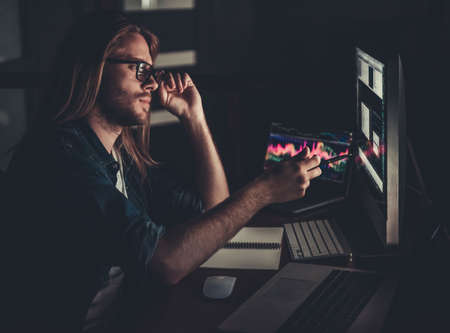 Handsome young businessman with shoulder-length blond hair and in casual wear and eyeglasses is working with a computer at nightの写真素材
