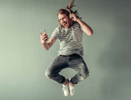 Stylish young man with shoulder-length blond hair and in headphones is listening to music while jumping, on gray backgroundの写真素材
