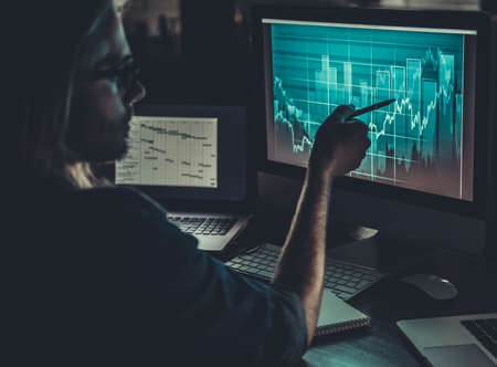 Handsome young businessman with shoulder-length blond hair and in casual wear and eyeglasses is studying graphs while working with a computer at nightの写真素材