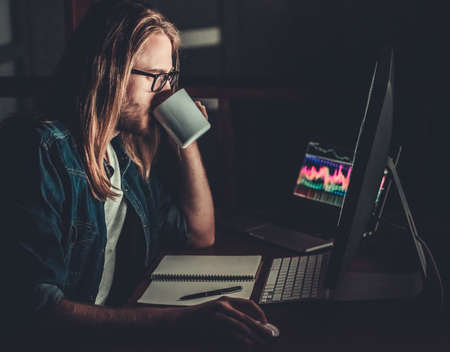 Handsome young businessman with shoulder-length blond hair and in eyeglasses is drinking coffee while working with a computer at nightの写真素材