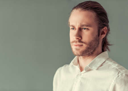 Portrait of stylish young businessman with shoulder-length blond hair and in white shirt, on gray backgroundの写真素材